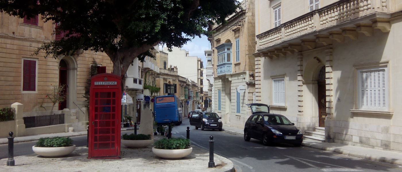 Das Bild einer Straße in der ein Bus für den Flughafentransfer steht. Daneben ist ein typisches Auto aus Malta mit dem Lenkrad auf der rechten Seite.