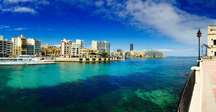 Das türkisblaue Meereswasser, gesehen von der nahen Küstenpromenade im Norden der Insel Malta. Der Blick geht auf das Meer und die auf die andere Seite der Bucht gelegenen Häuser. 