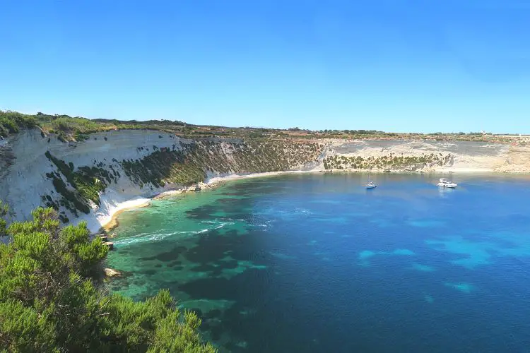 Die Bucht Il-Ħofra ż-Żgħira ist vom einer steilen Klippe umgeben. Auf dem Bild sieht man unterhalb der Klippen einen schmalen Sandstrand. Das Meer ist türkisblau gefäbrt, in der Bucht liegt eine Yacht vor Anker. 