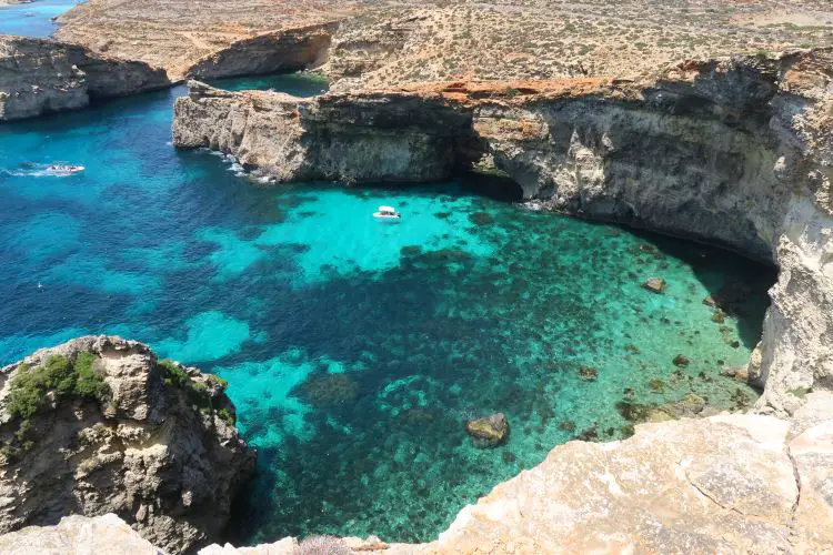 Der Blick von den Klippen auf Comino auf das türkislbaue Meer darunter und ein Schiff, dass in Richtung eines Felsenbogens fährt.