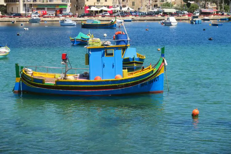 Ein blau-gelb-rotes Luzzu-Boot liegt in der türkislbauen Hafenbucht von Marsaskala vor Anker. 