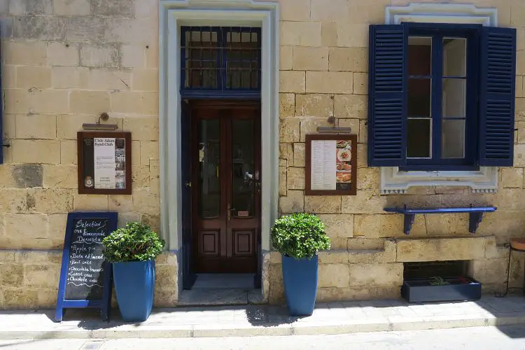 Die aus sandfarbenen Backseinen bestehende Wand eines Restaurants, mit einer kastanienfarbenen Tür und Blauen Fensterläden. 