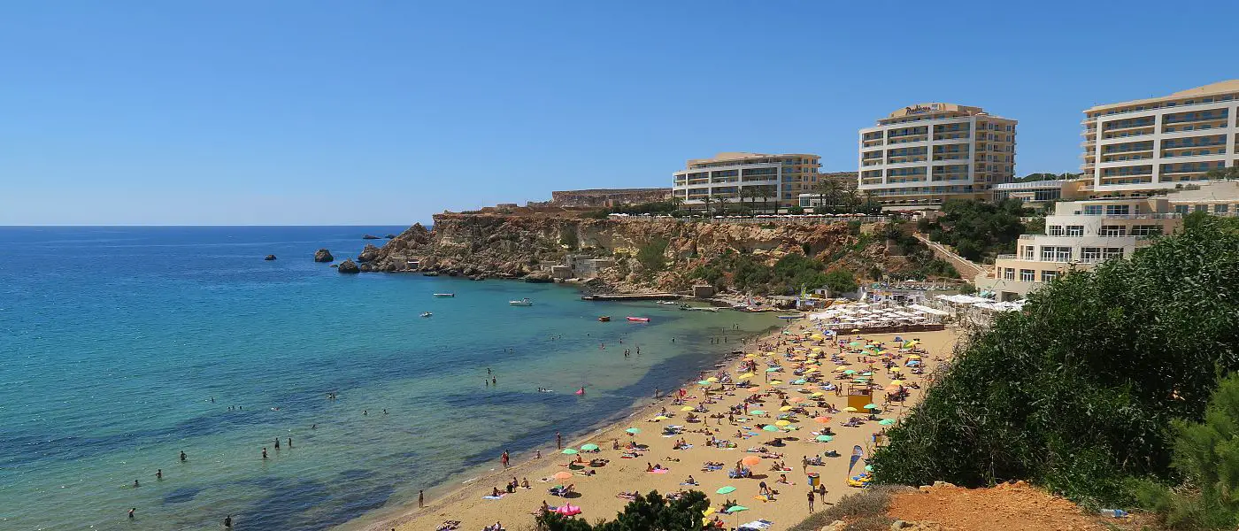 Das sommerliche, sonnige Wetter am langen Sandstrand der Golden Bay mit dem nahen Hotel im Juli. Davor ist das türkisblaue Meer an der Küste von Malta zu sehen.