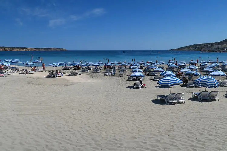 Der lange, breite Sandstrand mit Sonnenliegen und Sonnenschirmen in der Mellieha Bay. Der Blick geht auf das ruhige, türkisblaue Meer hinaus.