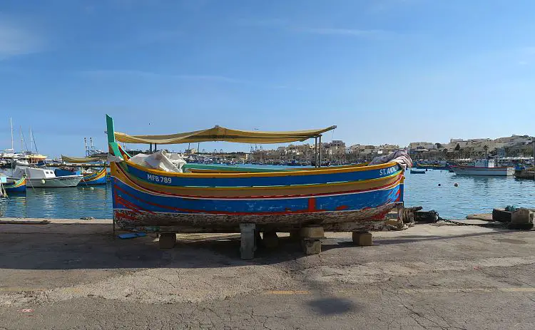 Ein rot-gelb-blaues Luzzu Boot steht am Land, neben dem Fischereihafen von Marsaxlokk.