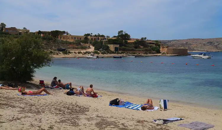 Ein Sandstrand mit Liegestühlen und Badegästen an einer schönen, türkisblauen Bucht mit einer Feriensiedlung im Hintergrund.